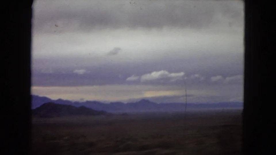 1967 Arizona home-movie frame of a desert plain and distant mountain skyline under heavy purple-gray clouds, partially framed by the vehicle interior.