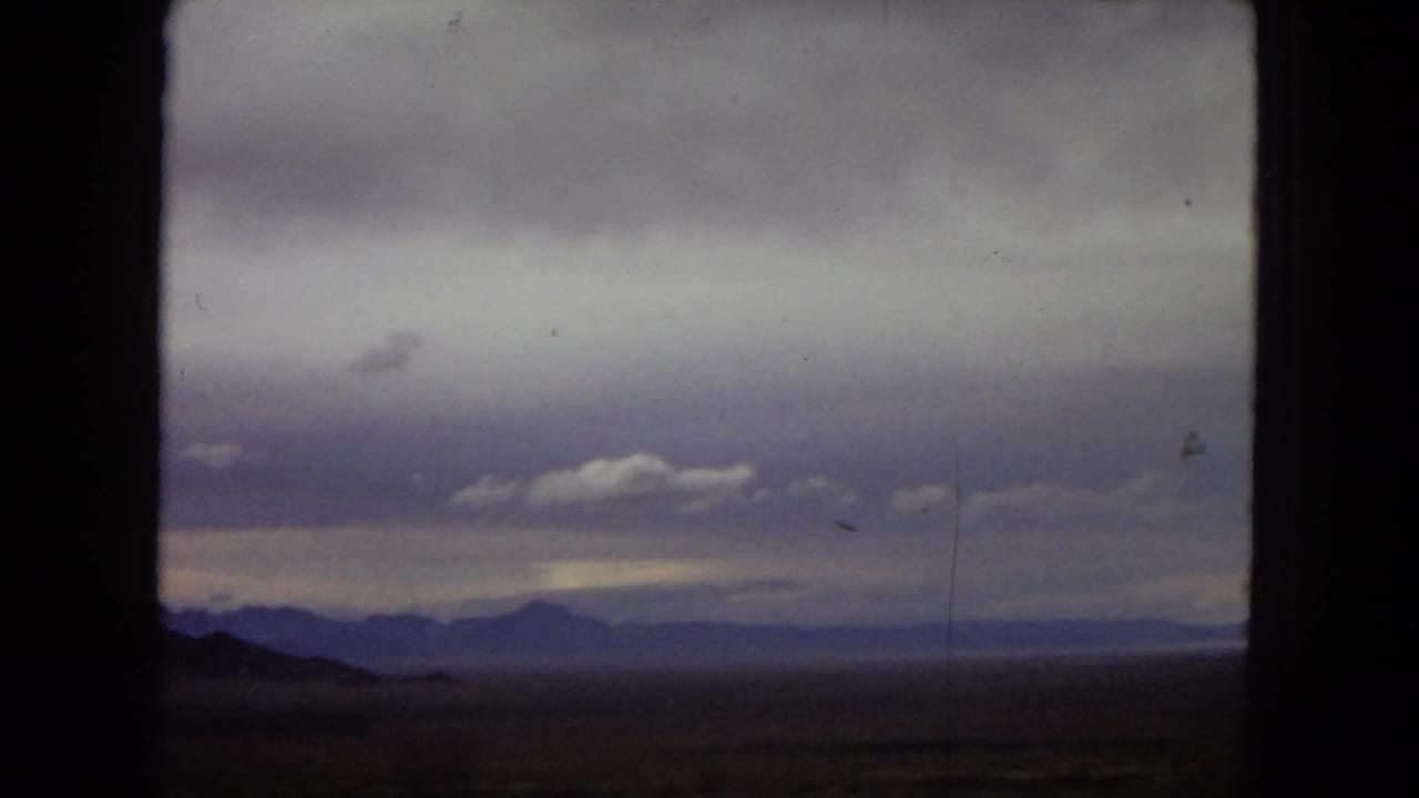 1967 Arizona home-movie frame showing open desert and distant mountain ridges through the dark silhouette of a car window beneath purple-gray clouds.