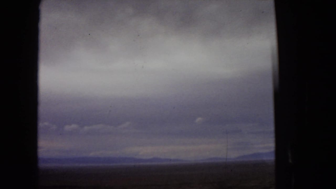 1967 Arizona travel footage frame with a flat desert horizon and far mountains viewed through a dark vehicle window under heavy, muted clouds.