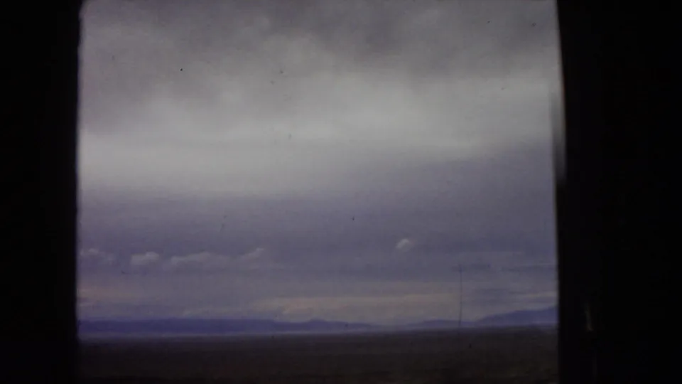 1967 Arizona travel footage frame with a flat desert horizon and far mountains viewed through a dark vehicle window under heavy, muted clouds.