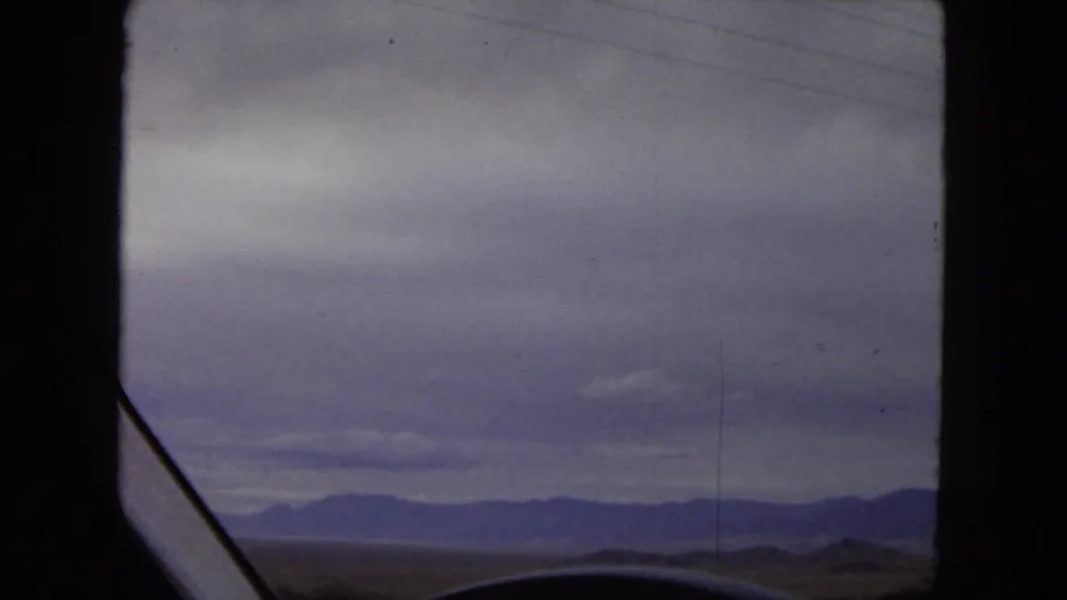 1967 Arizona 8mm frame showing a low desert foreground, distant mountains, and a broad overcast sky through the interior framing of a vehicle window.