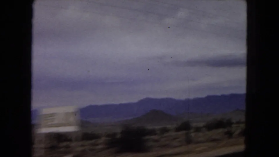 1967 Arizona home-movie frame with motion-blurred roadside desert, shrubs, and distant mountains seen from a moving vehicle beneath a heavy cloud layer.