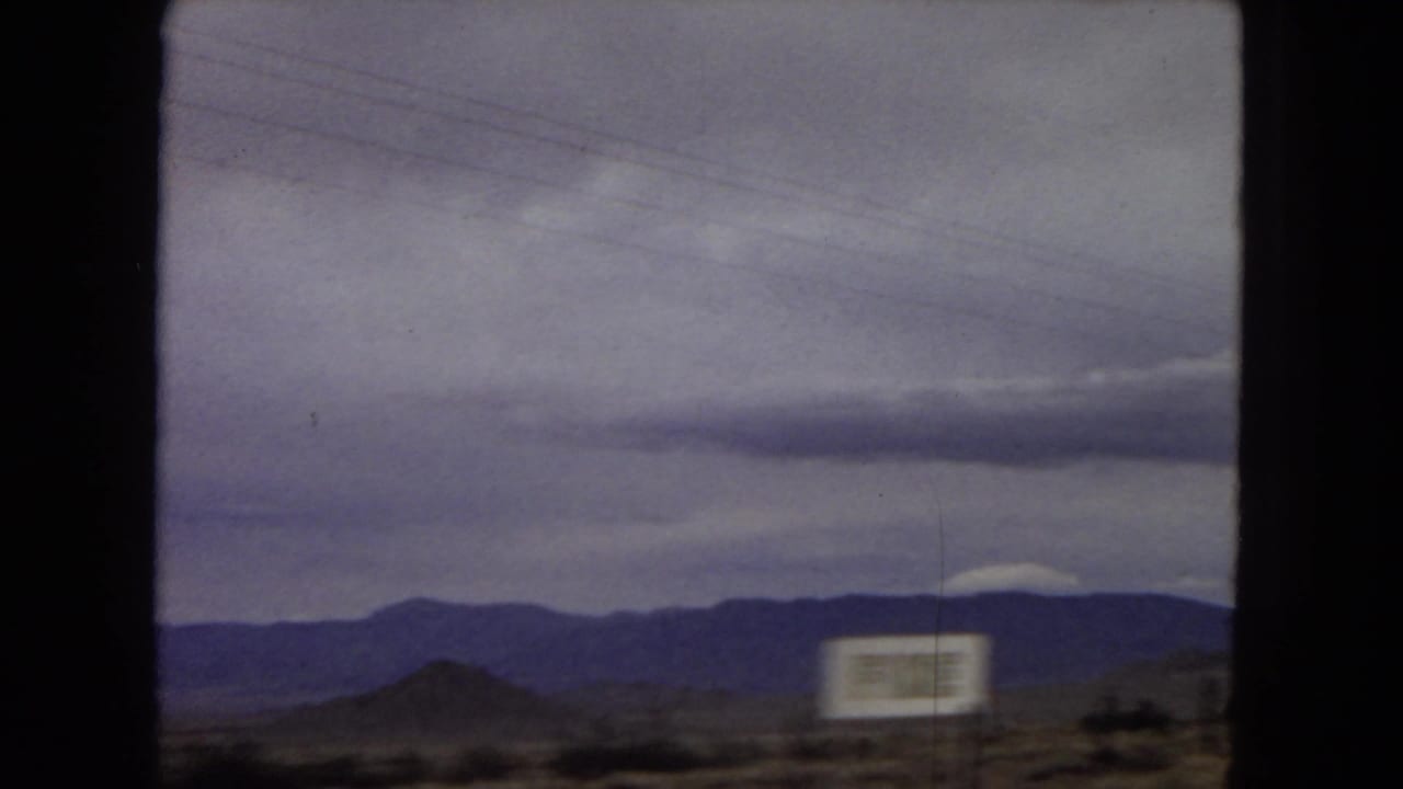 1967 Arizona road-travel frame showing distant desert mountains beneath a cloudy sky, with a blurred roadside sign and dark window framing from inside a moving vehicle.