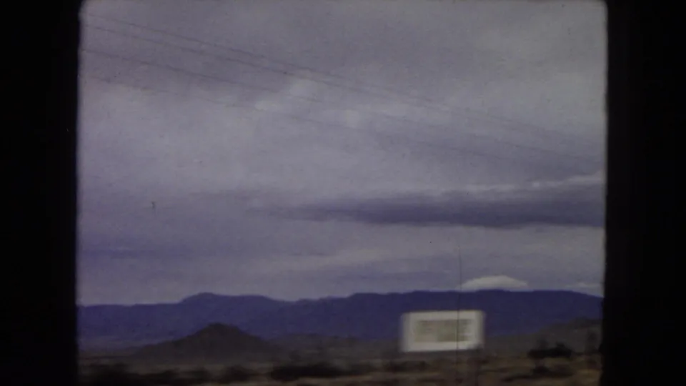 1967 Arizona road-travel frame showing distant desert mountains beneath a cloudy sky, with a blurred roadside sign and dark window framing from inside a moving vehicle.