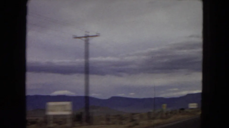 1967 Arizona highway view from a moving vehicle, with a utility pole, blurred roadside signs, distant mountains, and overcast desert sky.