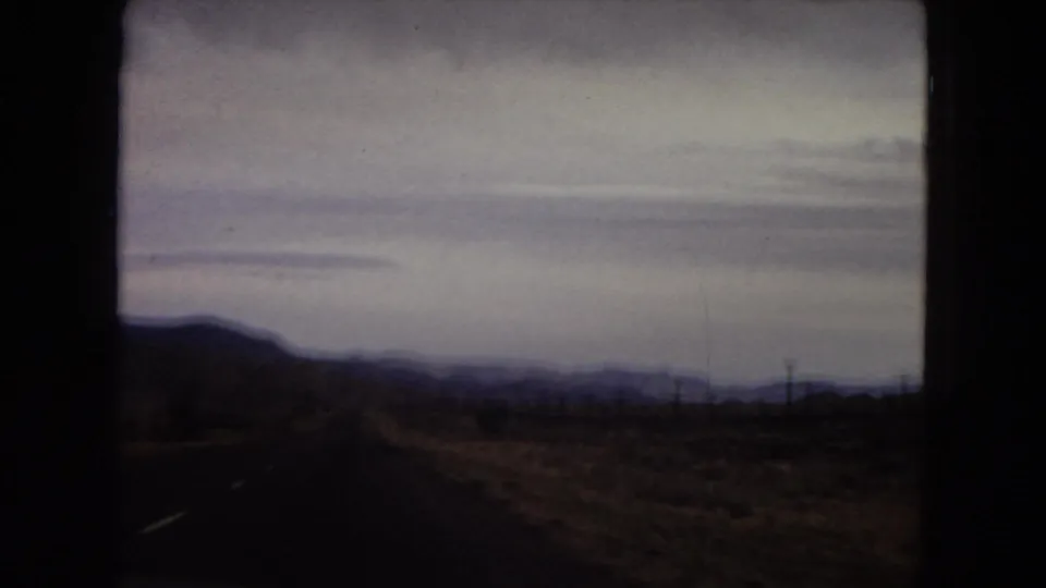 1967 Arizona in-vehicle road footage looking across dark desert foreground toward low mountains and utility lines under a cloudy sky.