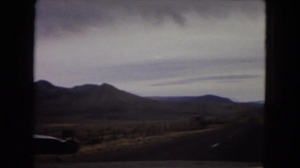 1967 Arizona road-trip frame with a paved road edge curving past desert hills and open scrubland beneath a cloudy sky, filmed from inside a vehicle.