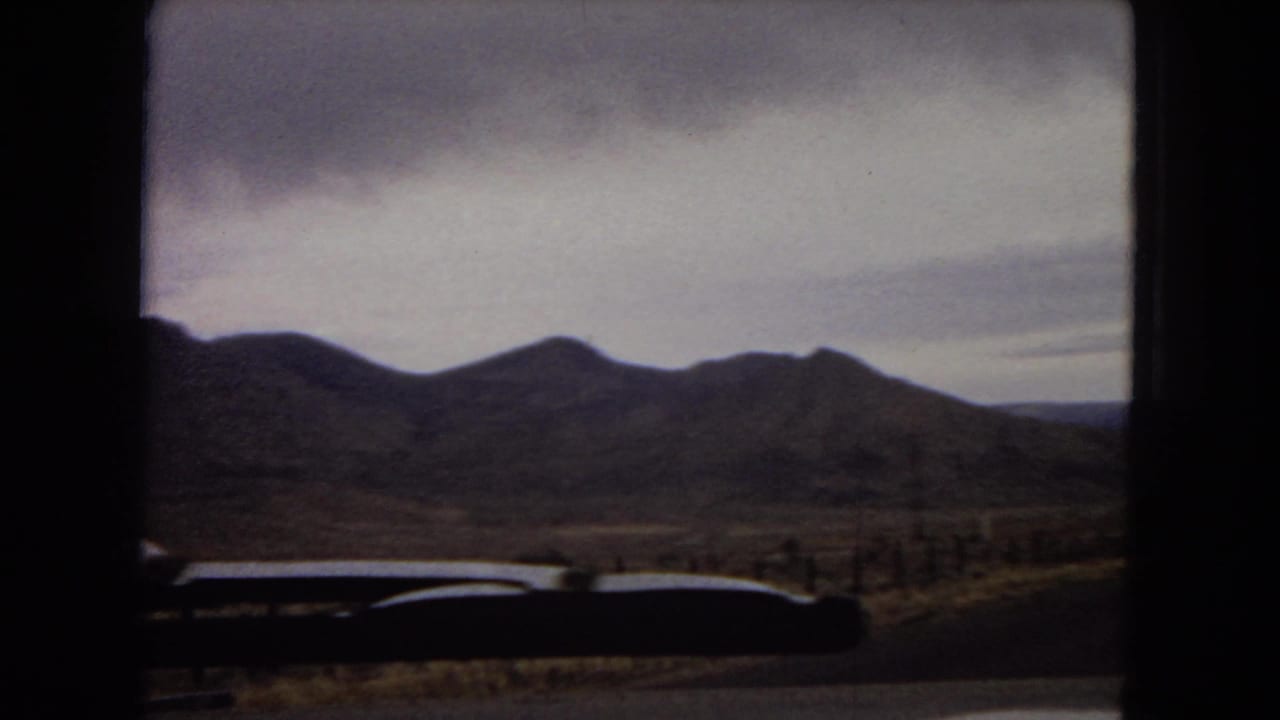 1967 Arizona home-movie frame from inside a moving car, showing a road curving through desert hills beneath a cloudy sky.