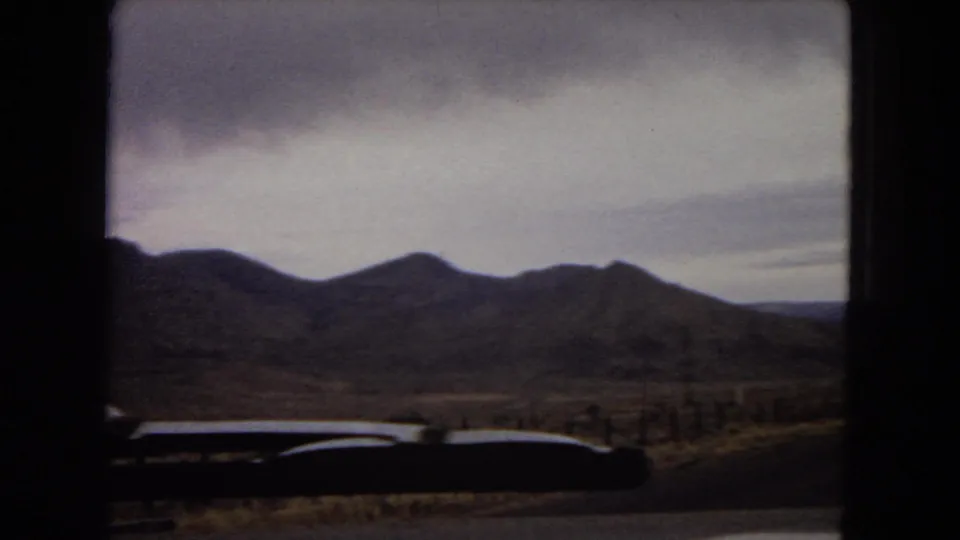 1967 Arizona home-movie frame from inside a moving car, showing a road curving through desert hills beneath a cloudy sky.
