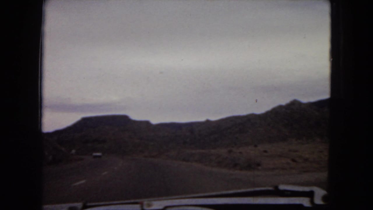 1967 Arizona road footage from a passenger perspective, with a distant vehicle on a highway between low mesas and rocky desert slopes.