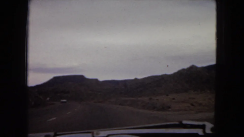 1967 Arizona road footage from a passenger perspective, with a distant vehicle on a highway between low mesas and rocky desert slopes.