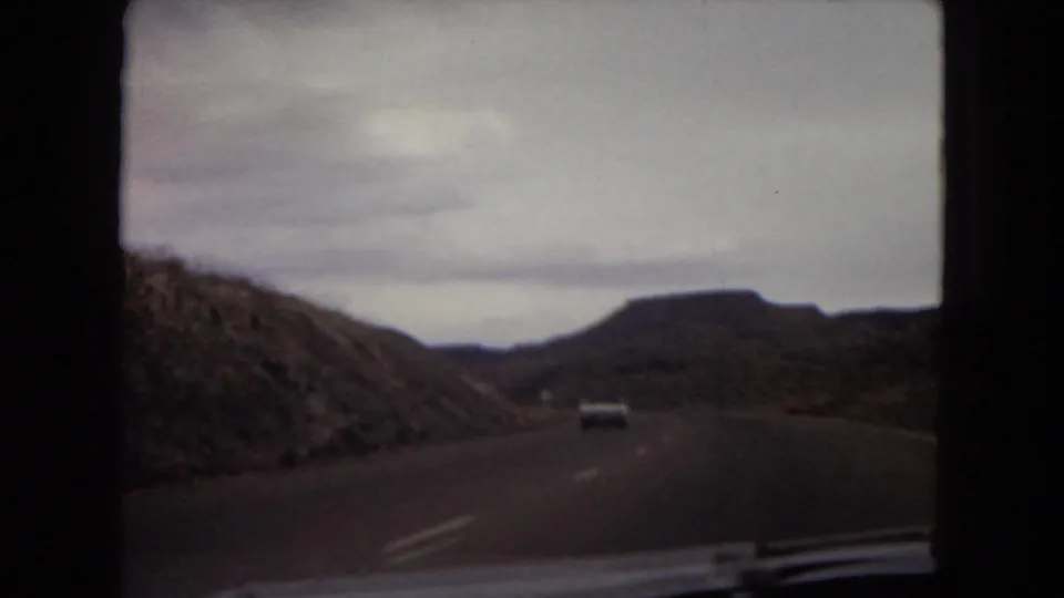 Passenger-shot 8mm frame of a highway cutting between rocky desert slopes, with another car visible ahead in the lane.