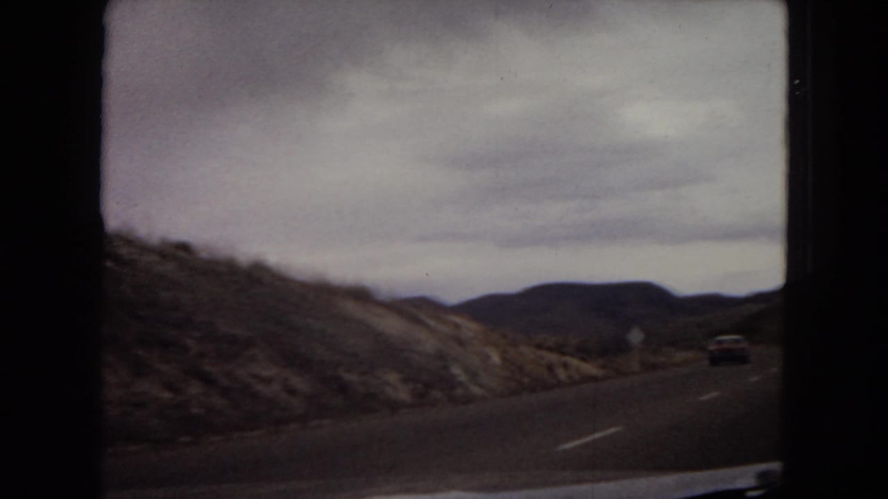 1967 8mm home-movie view from a moving car on a two-lane road through arid Arizona hills, with a distant vehicle and overcast sky.