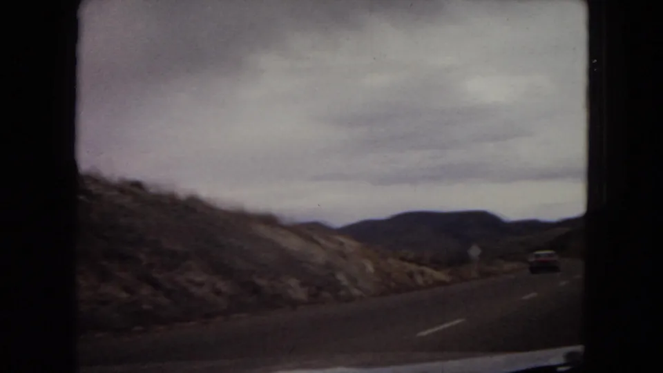1967 8mm home-movie view from a moving car on a two-lane road through arid Arizona hills, with a distant vehicle and overcast sky.