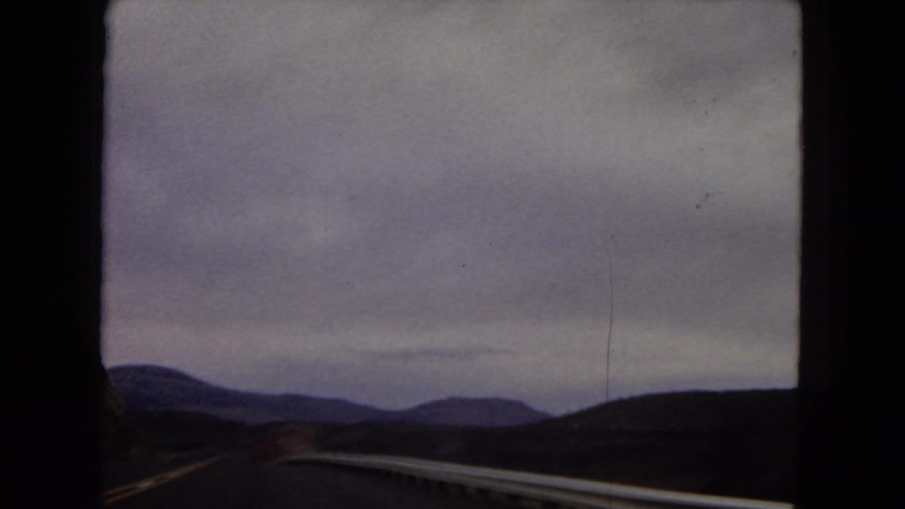 Passenger-side 8mm view across a roadside guardrail toward low Arizona mountain ranges beneath a dim, overcast sky.