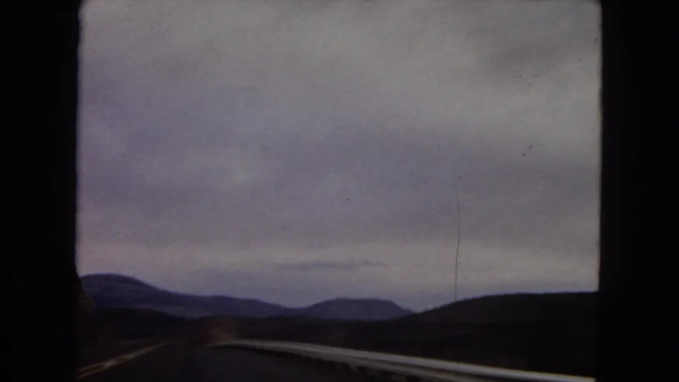 Passenger-side 8mm view across a roadside guardrail toward low Arizona mountain ranges beneath a dim, overcast sky.