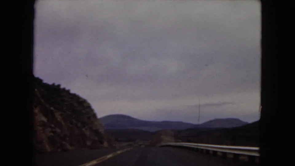 Vintage 8mm car-window view of a curving road bordered by a rocky desert slope, guardrail, and distant Arizona hills.