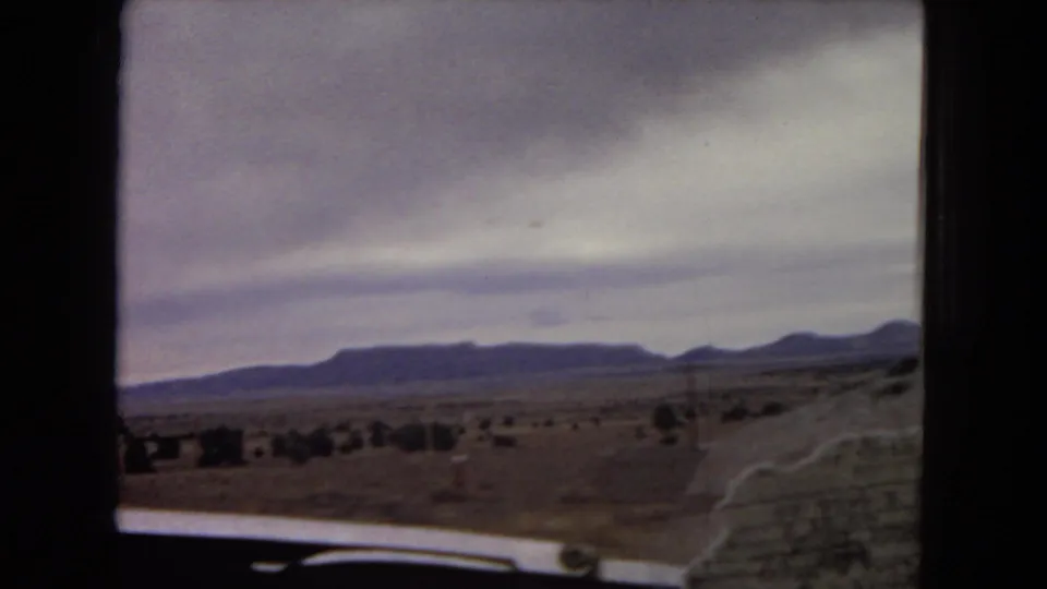 1967 8mm home-movie view across open Arizona desert toward distant mesa-like hills, framed by the moving car with part of the interior visible.