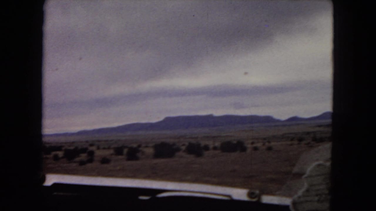1967 Arizona home-movie frame showing open desert, scattered shrubs, distant low mountains, and the edge of a car hood from a moving vehicle.