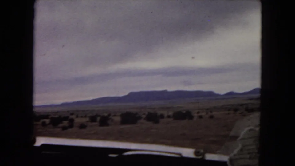 1967 Arizona home-movie frame showing open desert, scattered shrubs, distant low mountains, and the edge of a car hood from a moving vehicle.
