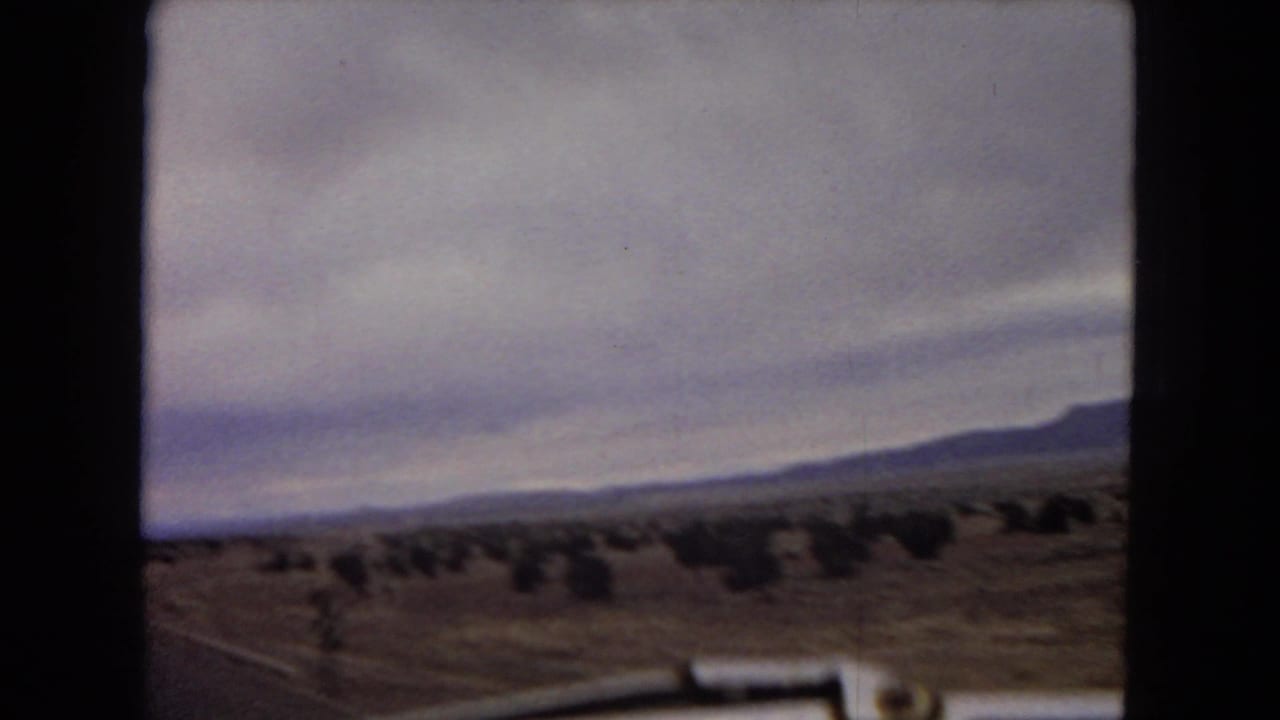 1967 Arizona 8mm frame with a soft, moving-car view across desert scrub toward low mountains beneath an overcast sky.