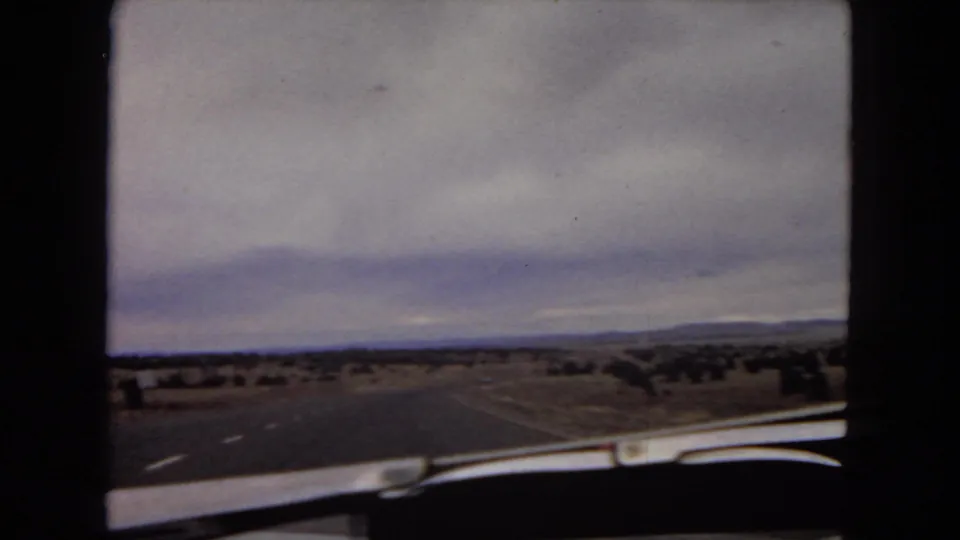 1967 Arizona road-trip frame showing a paved highway, desert shoulder, distant hills, and the car dashboard edge in the foreground.