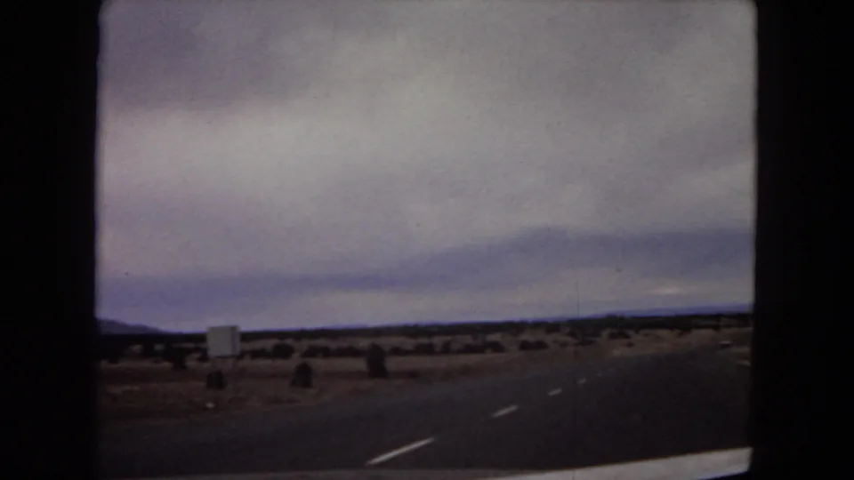 1967 Arizona home-movie frame showing a desert highway, small roadside sign, scattered shrubs, and distant mountains seen from a moving car.