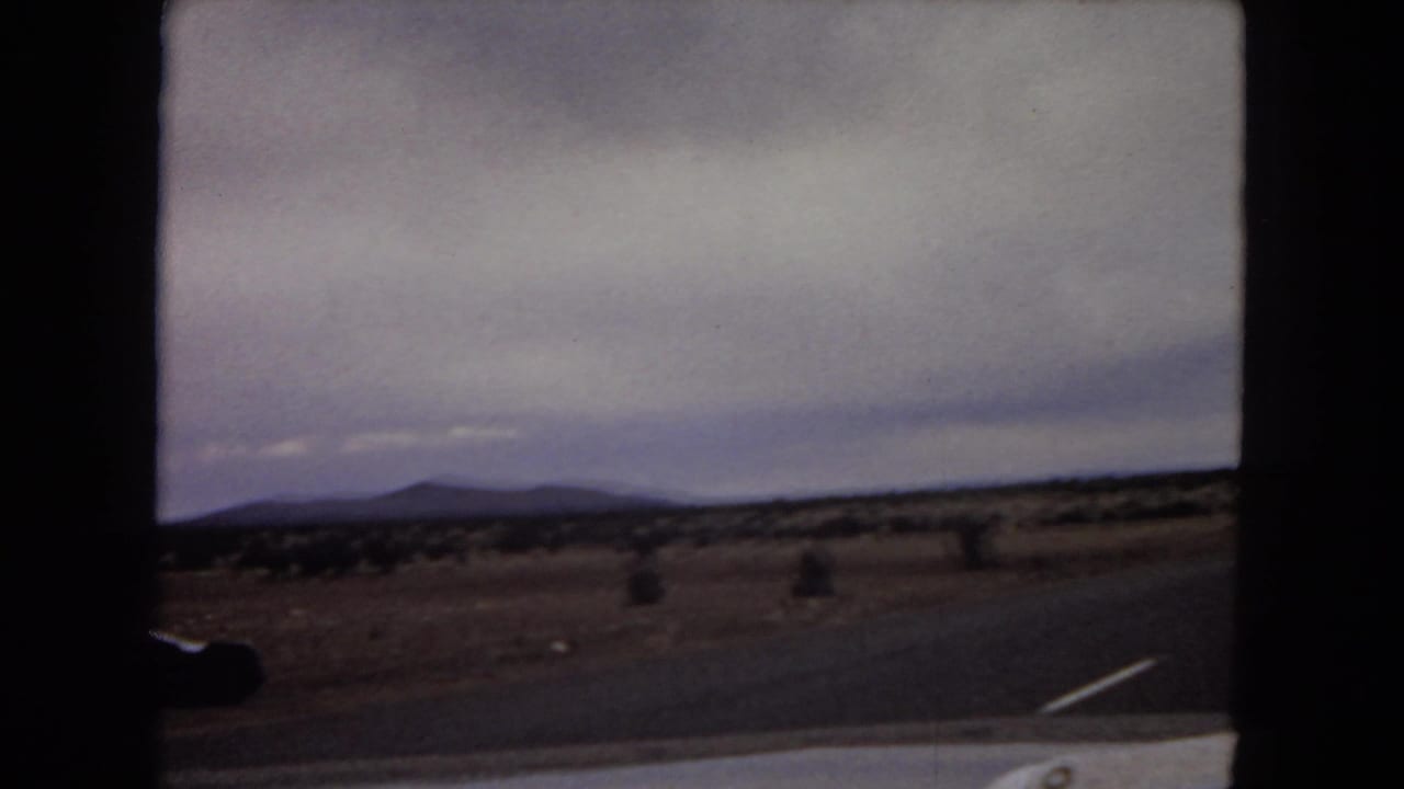 1967 Arizona home-movie frame showing scrubby open grassland, distant mountain ridges, cloudy sky, and a dark car-window edge from a moving vehicle.