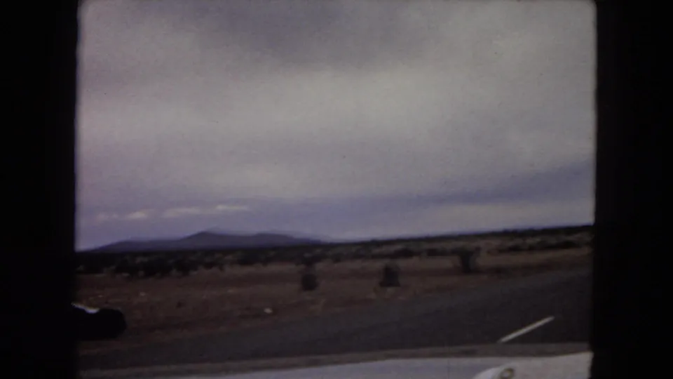 1967 Arizona home-movie frame showing scrubby open grassland, distant mountain ridges, cloudy sky, and a dark car-window edge from a moving vehicle.