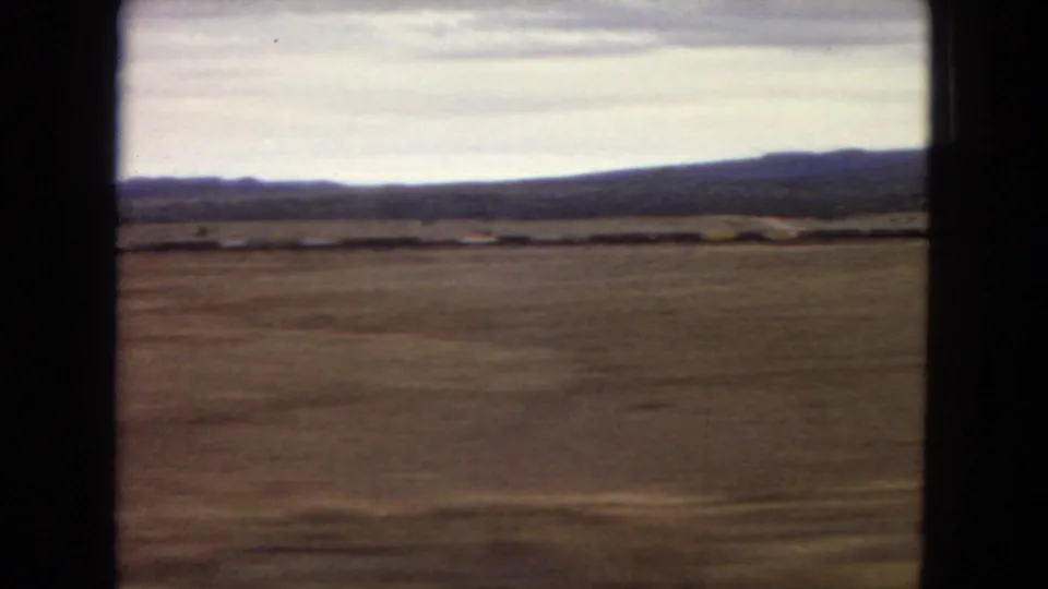 1967 Arizona home-movie frame showing a broad dry plain, low brush, a dark mountain ridge, and pale cloud cover from a moving vehicle perspective.