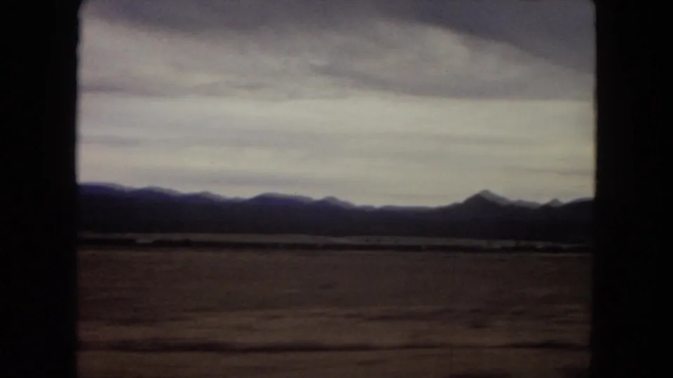 1967 Arizona 8mm frame with a flat scrubby foreground, distant mountain silhouettes, layered cloudy sky, and dark film-frame edges.