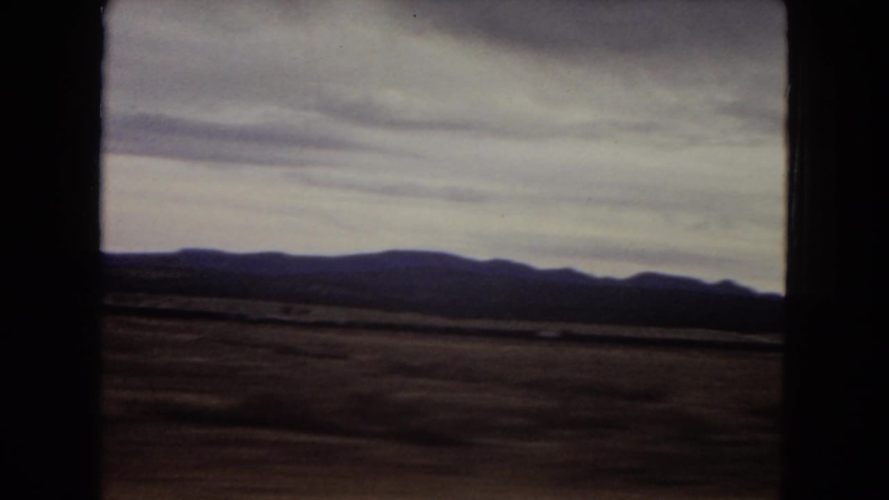 1967 Arizona home-movie view of a broad desert plain, low mountains, cloudy sky, and dark car-window framing from a moving vehicle.