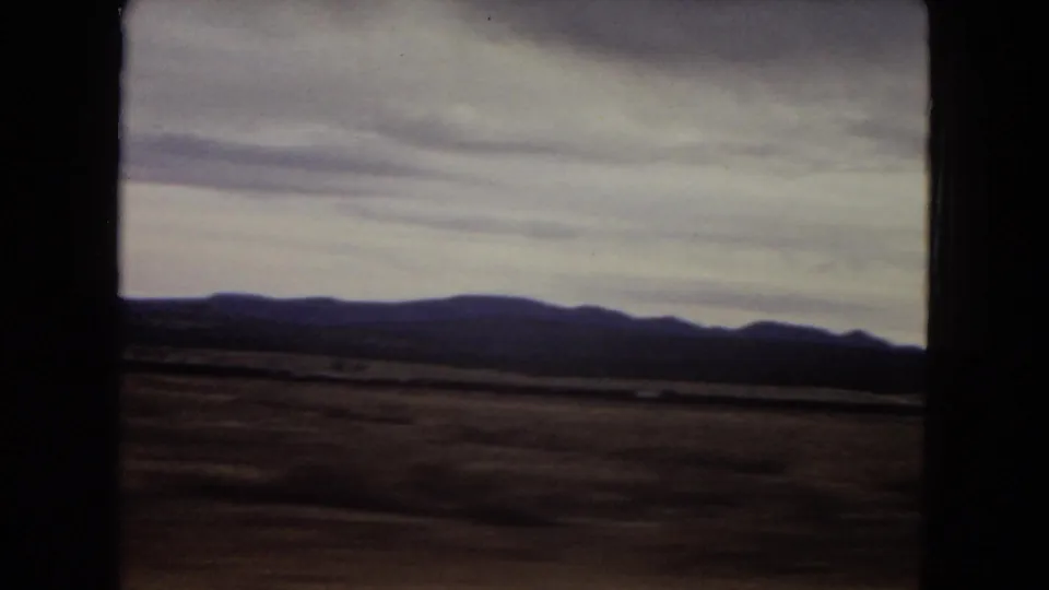 1967 Arizona home-movie view of a broad desert plain, low mountains, cloudy sky, and dark car-window framing from a moving vehicle.