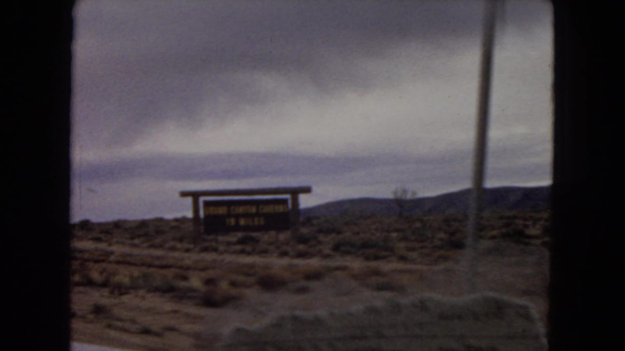 A wooden roadside sign appears beside scrubby desert terrain and distant hills during the 1967 Arizona car-window travel footage.