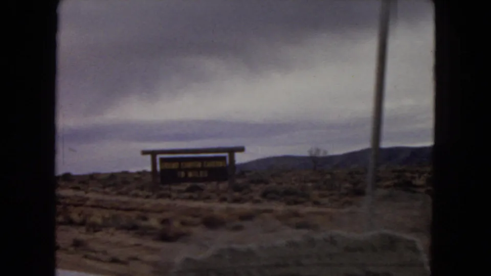 A wooden roadside sign appears beside scrubby desert terrain and distant hills during the 1967 Arizona car-window travel footage.