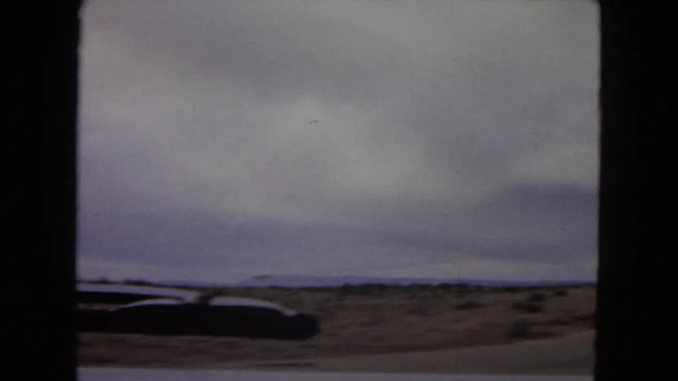 Open Arizona desert, distant low hills, cloudy sky, and a dark edge of the car appear in this 1967 moving-vehicle home-movie frame.