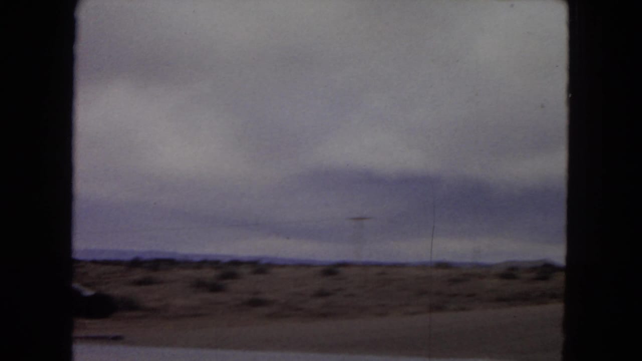 1967 Arizona home-movie frame showing a flat desert landscape with sparse scrub, distant low horizon, and heavy cloud cover, viewed from a moving vehicle.
