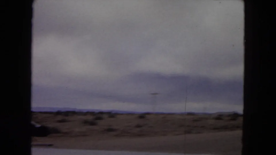 1967 Arizona home-movie frame showing a flat desert landscape with sparse scrub, distant low horizon, and heavy cloud cover, viewed from a moving vehicle.