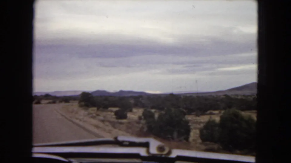 1967 Arizona road-trip view from inside a car, looking down a paved desert road toward low mountains beneath layered clouds.
