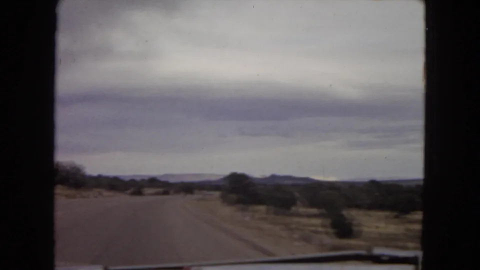1967 Arizona home-movie frame of a paved desert road viewed from a moving vehicle, with scrubland, low hills, and a cloudy sky ahead.