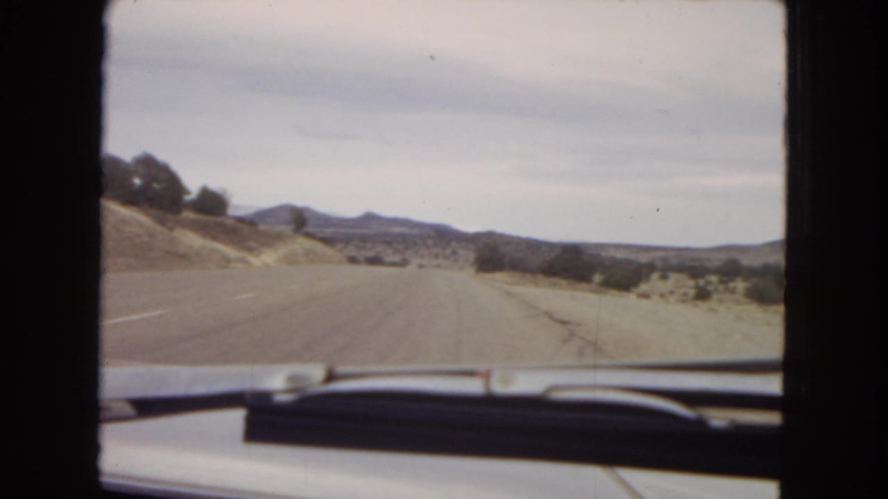 Driver’s-eye 8mm view from inside a car on a paved road through arid Arizona landscape, with low hills and distant mountains under a pale sky.