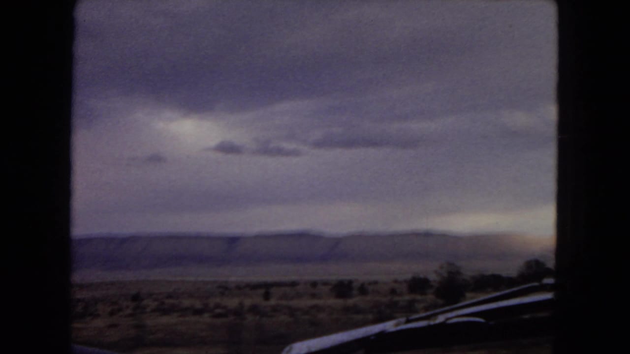 View from a moving car across scrubby desert and a distant ridge beneath heavy clouds, with part of the car visible at the lower right.