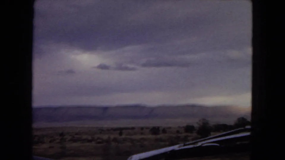 View from a moving car across scrubby desert and a distant ridge beneath heavy clouds, with part of the car visible at the lower right.