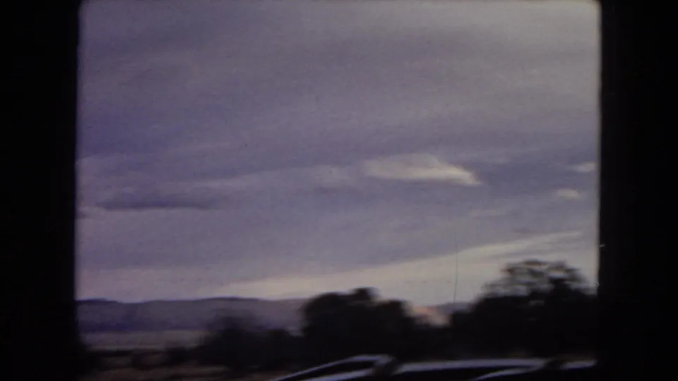 Motion-blurred 8mm roadside view with dark trees, low hills, and a broad cloudy sky during travel through the Southwest landscape.