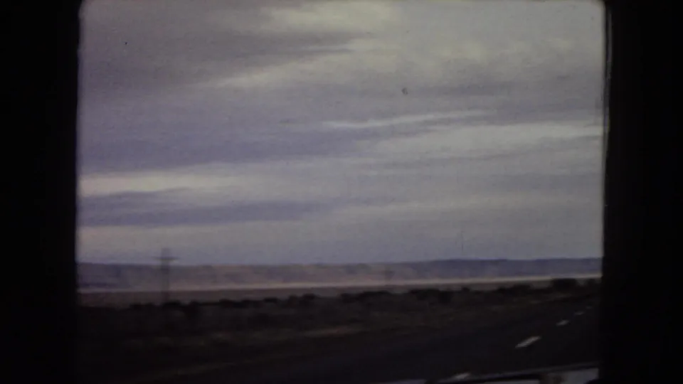 A moving car-window view shows roadside desert, a paved road edge with white lane markings, distant hills, and layered clouds over the Arizona landscape.