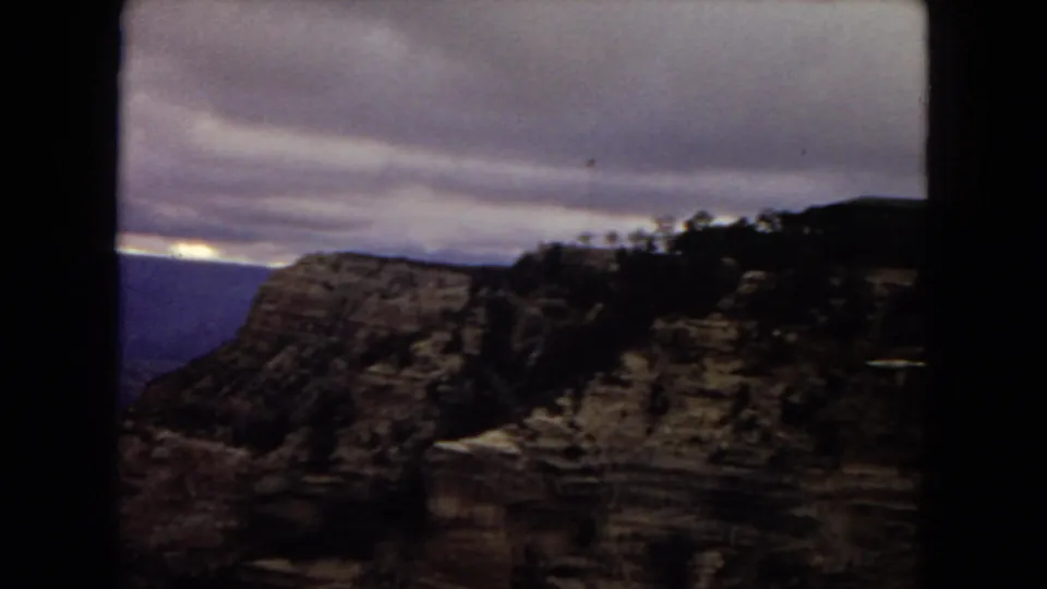 The footage shifts to a canyon overlook, with layered rock formations, a dark rim silhouette, sparse trees, and heavy clouds over the distant canyon landscape.