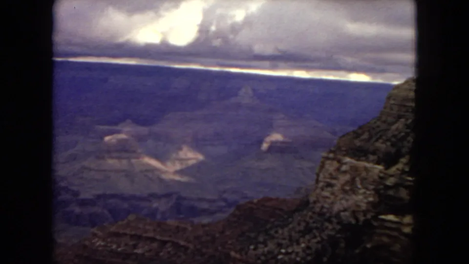 A Grand Canyon overlook appears in 1967 home-movie footage, showing a rocky foreground, deep canyon walls, distant layered formations, and a bright break in the cloud cover.