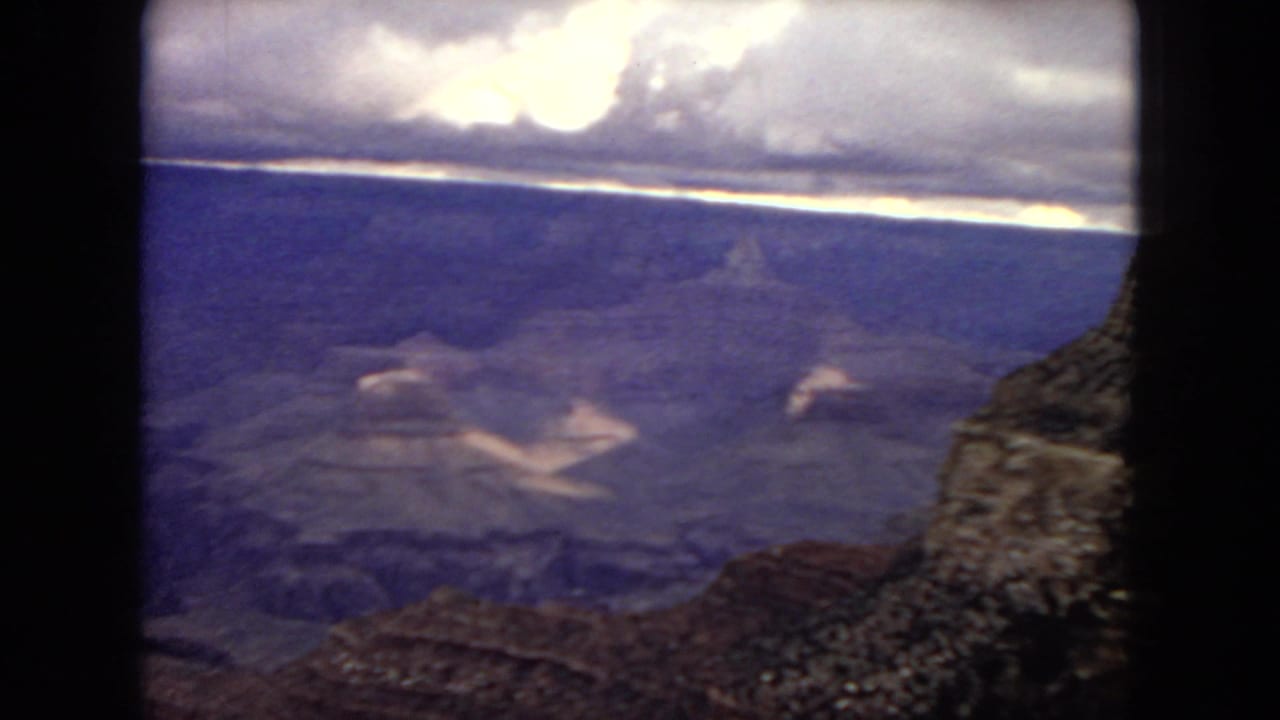 1967 8mm home-movie frame showing the Grand Canyon from a high rim viewpoint, with layered rock formations, a dark foreground edge, and bright cloud light above.