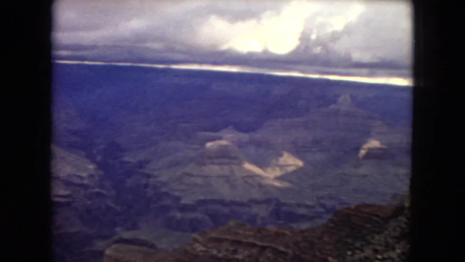 Wide 1967 Grand Canyon view in aged 8mm color, showing blue-purple canyon layers, distant horizon, bright clouds, and dark film borders.