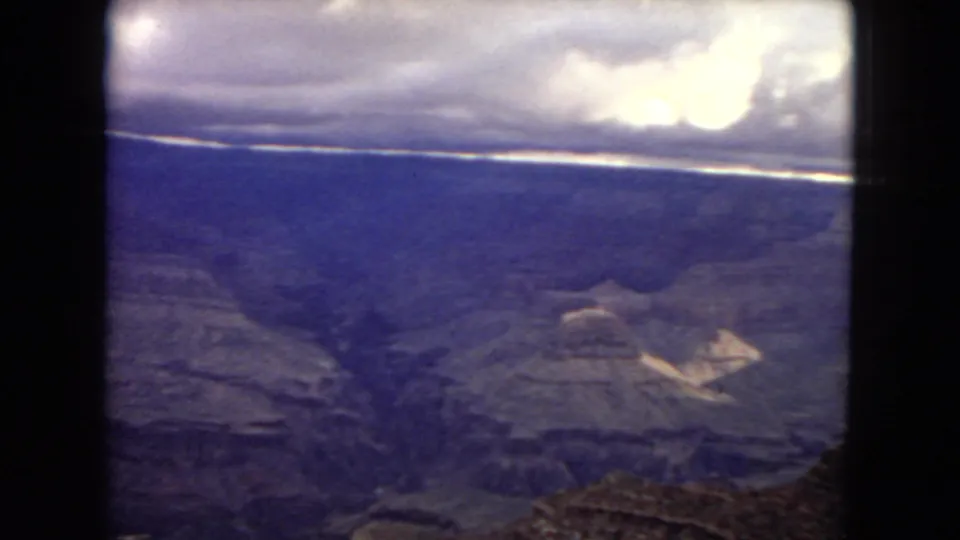 Home-movie frame of the Grand Canyon from the rim in 1967, with shadowed ravines, layered rock shelves, a cloudy sky, and visible 8mm gate edges.